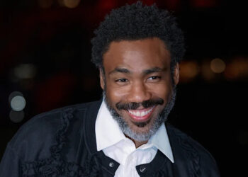 Donald Glover, smiling with a beard, wearing a white shirt and dark jacket, against a blurred dark background.