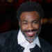 Donald Glover, smiling with a beard, wearing a white shirt and dark jacket, against a blurred dark background.