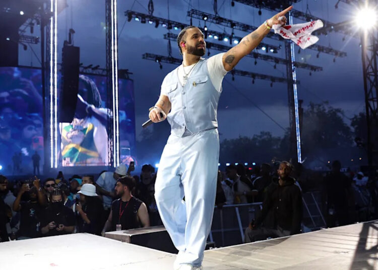 Drake performing on stage in a light blue outfit, holding a microphone and pointing to the crowd, with stage lights behind him.