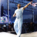 Drake performing on stage in a light blue outfit, holding a microphone and pointing to the crowd, with stage lights behind him.