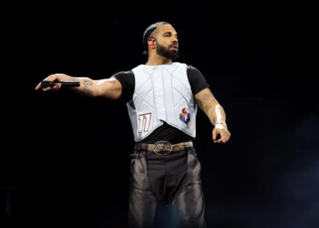 Drake performing on stage, wearing a white vest and black shirt, holding a microphone. He is getting ready to release 'Iceman' his latest album.