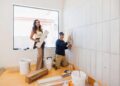 Two individuals tiling a wall in a room under renovation.