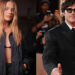 Margot Robbie in a black outfit poses confidently, and Jacob Elordi in a black suit and sunglasses gives a thumbs up at a formal event.