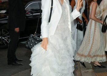 A person wearing a white layered dress and a blazer, holding a black handbag, walking on the street.