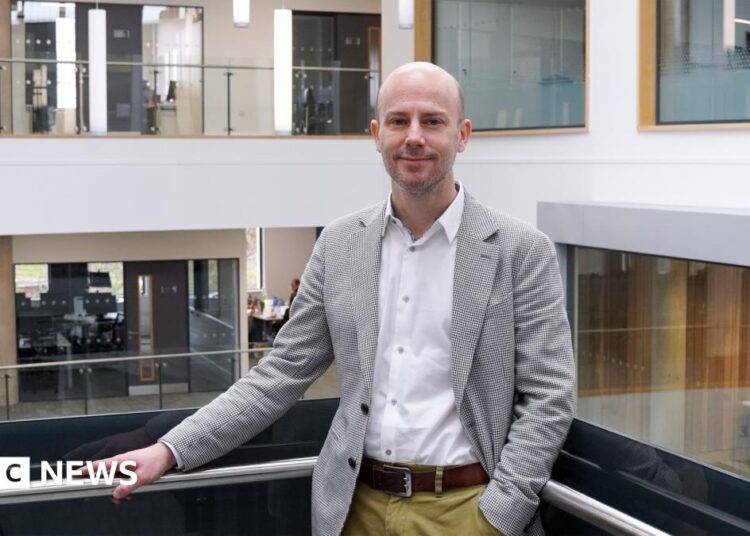 Andrew is wearing a black and white plaid blazer, a white shirt and chinos. He has short hair and is standing with his arm resting on a balcony. He is smiling at the camera. There are offices behind him.