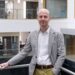 Andrew is wearing a black and white plaid blazer, a white shirt and chinos. He has short hair and is standing with his arm resting on a balcony. He is smiling at the camera. There are offices behind him.