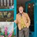 An elderly gentleman is standing in front of a vibrant blue entrance to a café, dressed in a tan jacket and gray trousers. The café’s exterior is adorned with colorful flowers and has a glass door with the word "CAFE" prominently displayed in illuminated letters.