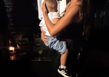 A woman holding a child in a dimly lit setting with a shirt displayed in the background.