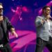 Two male singers stand on a large concert stage in front of a bright pink and purple LED screen. Both wear sunglasses and hold handheld microphones. One is dressed in a black jacket and black trousers, while the other wears a patterned shirt over a white top with black trousers and a chain at his waist. Stage lighting and part of a band setup are visible in the background.