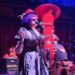 A woman with purple hair and a red hat sings into a microphone while a musician behind her plays a red sousaphone on stage.