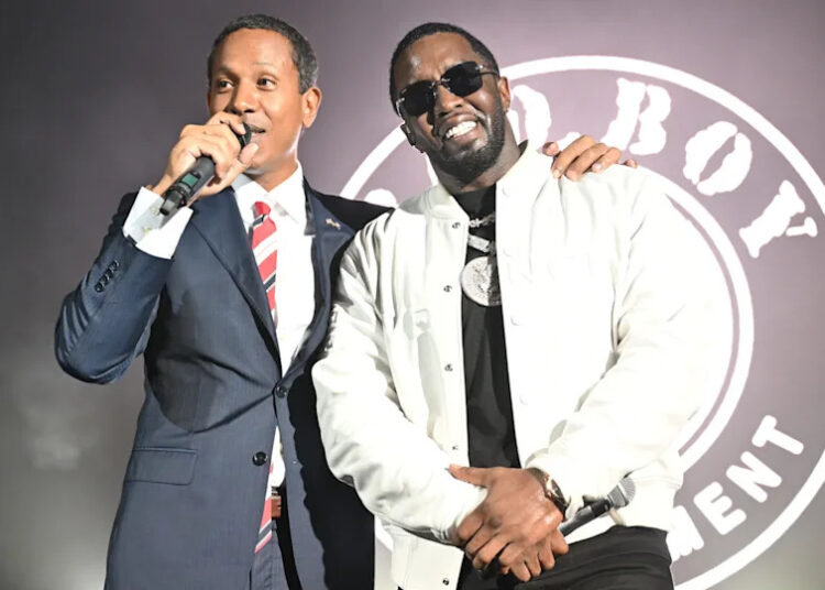 Shyne and Diddy stand together on stage, one in a suit holding a microphone, and the other in a white jacket and sunglasses.