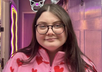 A woman with long brown hair and glasses in a pink top with hearts on, standing in a corridor lit with a pink light
