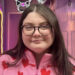 A woman with long brown hair and glasses in a pink top with hearts on, standing in a corridor lit with a pink light