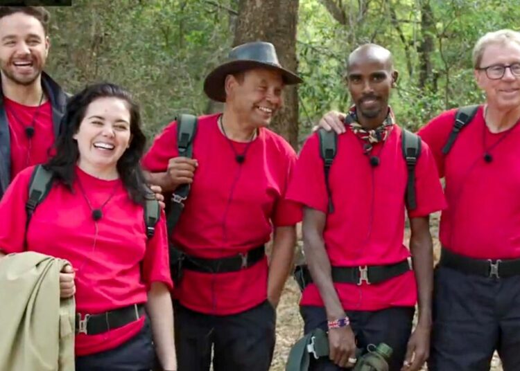 Adam Thomas, Scarlett Moffatt, Craig Charles, Mo Farah and Harry Redknapp standing together, smiling and all wearing red I'm A Celebrity T-shirts