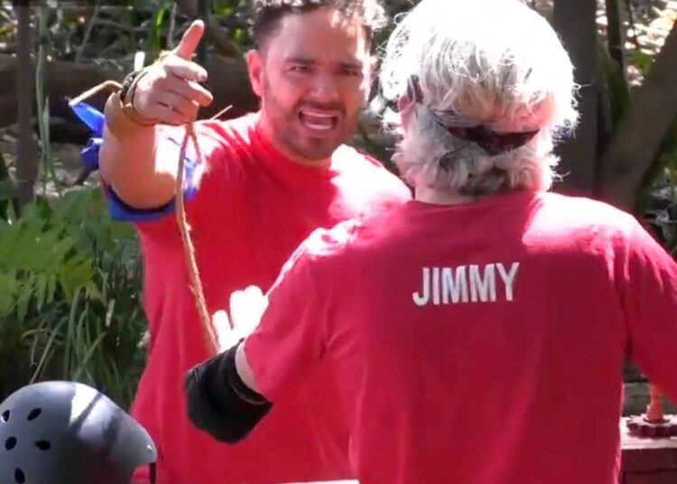 Two individuals dressed in red t-shirts, one with a visible "JIMMY" print on the back, are standing close together in an outdoor setting, with one of them gesturing with a pointed finger.