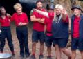 A group of individuals dressed in red shirts and blue shorts are gathered together in an outdoor setting. They appear to be engaged in an activity, with one person standing out by holding an object and gesturing while the others look on. The background reveals a rustic and natural environment with a rustic structure and trees.
