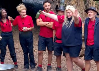 A group of individuals dressed in red shirts and blue shorts are gathered together in an outdoor setting. They appear to be engaged in an activity, with one person standing out by holding an object and gesturing while the others look on. The background reveals a rustic and natural environment with a rustic structure and trees.