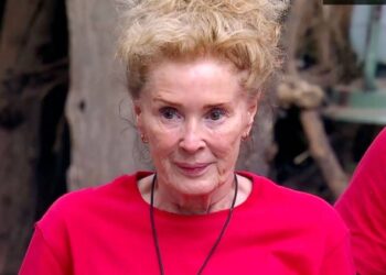An elderly woman with short blonde hair is wearing a red shirt and a necklace. She is standing outside, smiling at the camera, with a tree trunk and a partially visible person in a red shirt behind her.