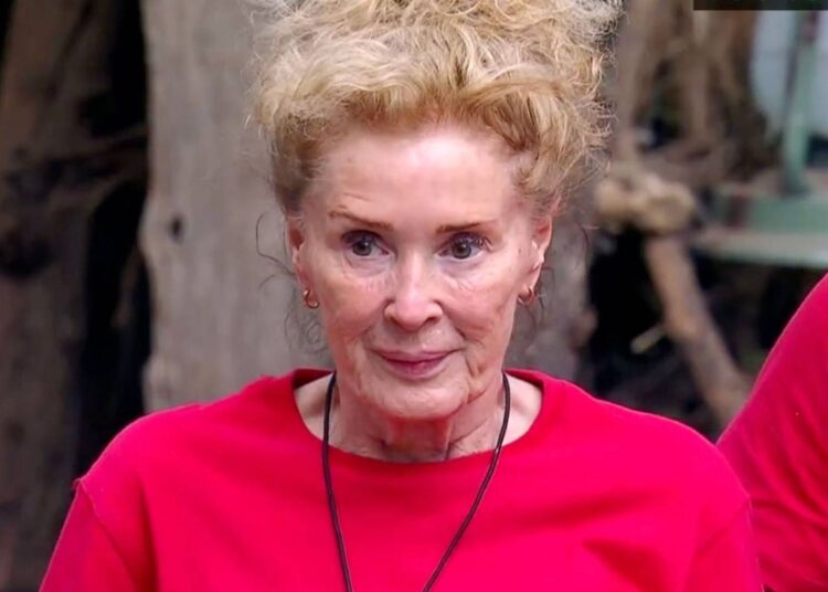 An elderly woman with short blonde hair is wearing a red shirt and a necklace. She is standing outside, smiling at the camera, with a tree trunk and a partially visible person in a red shirt behind her.