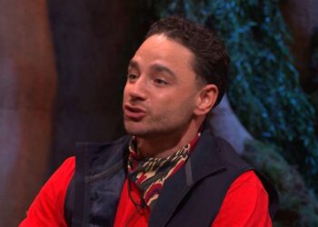 An individual dressed in a red long-sleeve shirt and a black vest with a patterned scarf is speaking on stage. The background is blurred with green and white colors.