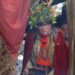 An individual adorned in a red robe and a garland of greenery is peering through a curtain embellished with red fabric.