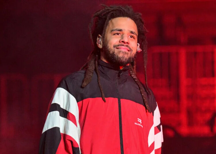 J. Cole performing on stage, wearing a red and black jacket, with a confident expression and dreadlocks.