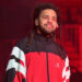 J. Cole performing on stage, wearing a red and black jacket, with a confident expression and dreadlocks.