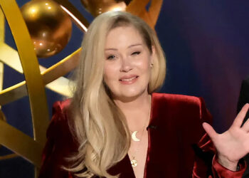 Christina Applegate, in a red outfit, gestures while speaking, with a golden decorative background behind her.