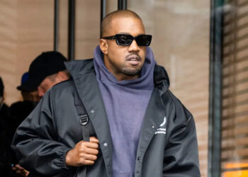Kanye West wearing sunglasses and a dark jacket over a hoodie, walking outdoors.