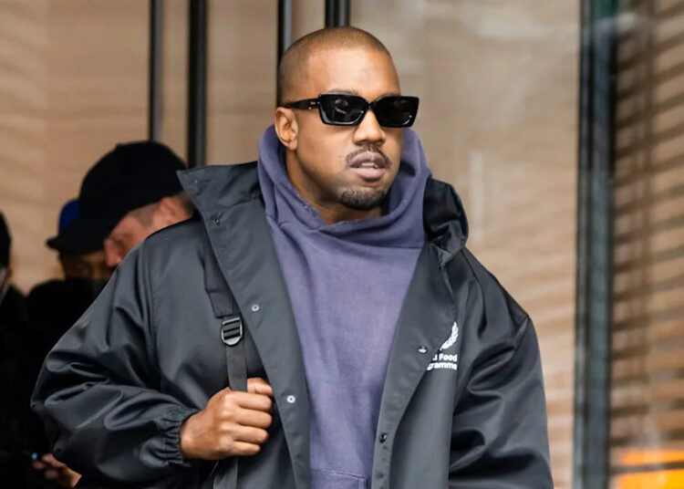 Kanye West wearing sunglasses and a dark jacket over a hoodie, walking outdoors.