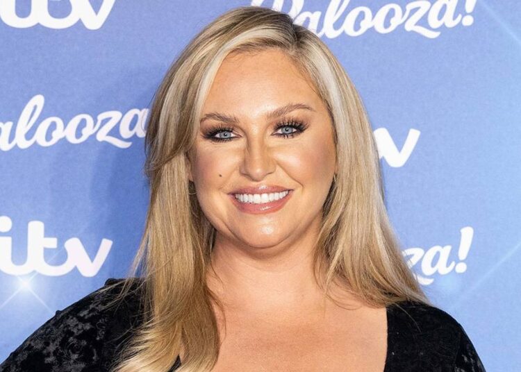 An adult woman with blonde hair is standing against a blue backdrop, smiling, and dressed in a black outfit. The backdrop includes white text that reads "Palooza" and "ITV" in a slanted manner.