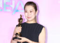 'KPop Demon Hunters' co-director Maggie Kang in a black dress holds a gold trophy, smiling against a pink background.