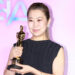 'KPop Demon Hunters' co-director Maggie Kang in a black dress holds a gold trophy, smiling against a pink background.