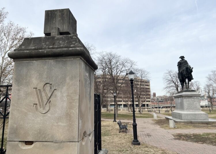 Outdoors photo on a cloudy day shows a park in wintertime. Trees are bare and there's a bronze statue of George Washington riding a horse on top of a marble pedestal. In the foreground is a concrete column that shows "WS" chiseled in it.