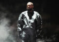Kanye West wearing a dark, oversized outfit, standing amidst smoke on stage. He just released his latest album, Bully.