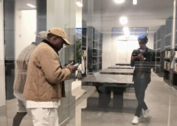 Ye and Nico Ballesteros are seen through a glass wall in a modern library or office. One is using a phone, while the other is reflected in the glass.