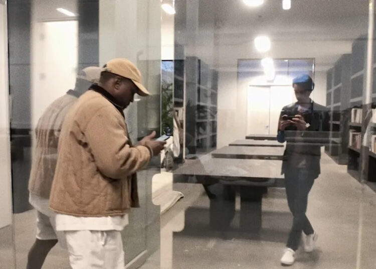 Ye and Nico Ballesteros are seen through a glass wall in a modern library or office. One is using a phone, while the other is reflected in the glass.