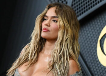 Karol. G with long, wavy hair poses confidently in front of a textured wall.