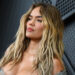Karol. G with long, wavy hair poses confidently in front of a textured wall.