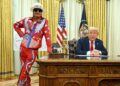 In a formal setting, a man in a red and blue costume stands beside an individual in a suit seated at a desk, with American flags
