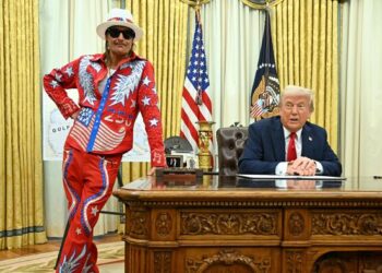 In a formal setting, a man in a red and blue costume stands beside an individual in a suit seated at a desk, with American flags