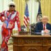 In a formal setting, a man in a red and blue costume stands beside an individual in a suit seated at a desk, with American flags