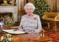 Queen Elizabeth II sits at a desk in  a grand drawing room with a Christmas tree behind her