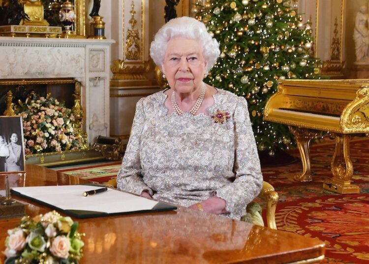 Queen Elizabeth II sits at a desk in  a grand drawing room with a Christmas tree behind her