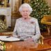 Queen Elizabeth II sits at a desk in  a grand drawing room with a Christmas tree behind her