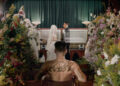 The cover art for Yeat's 'ADL' album. The art depicts a shirtless man with tattoos sitting in a chapel, facing a coffin and a couple in wedding attire. Surrounded by floral arrangements.