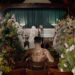 The cover art for Yeat's 'ADL' album. The art depicts a shirtless man with tattoos sitting in a chapel, facing a coffin and a couple in wedding attire. Surrounded by floral arrangements.