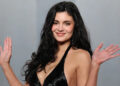 Kylie Jenner smiling, wearing a black dress, with her hands raised in a waving gesture.