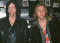Two musicians standing indoors with long hair and leather jackets