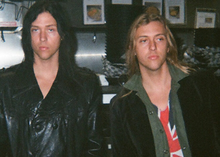 Two musicians standing indoors with long hair and leather jackets